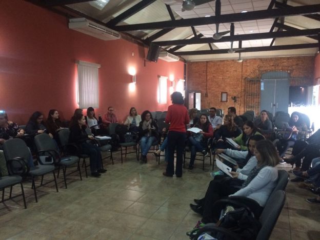 Descrição da imagem: foto de um grupo de pessoas sentadas em um semicírculo. À frente deles há uma mulher de pé.